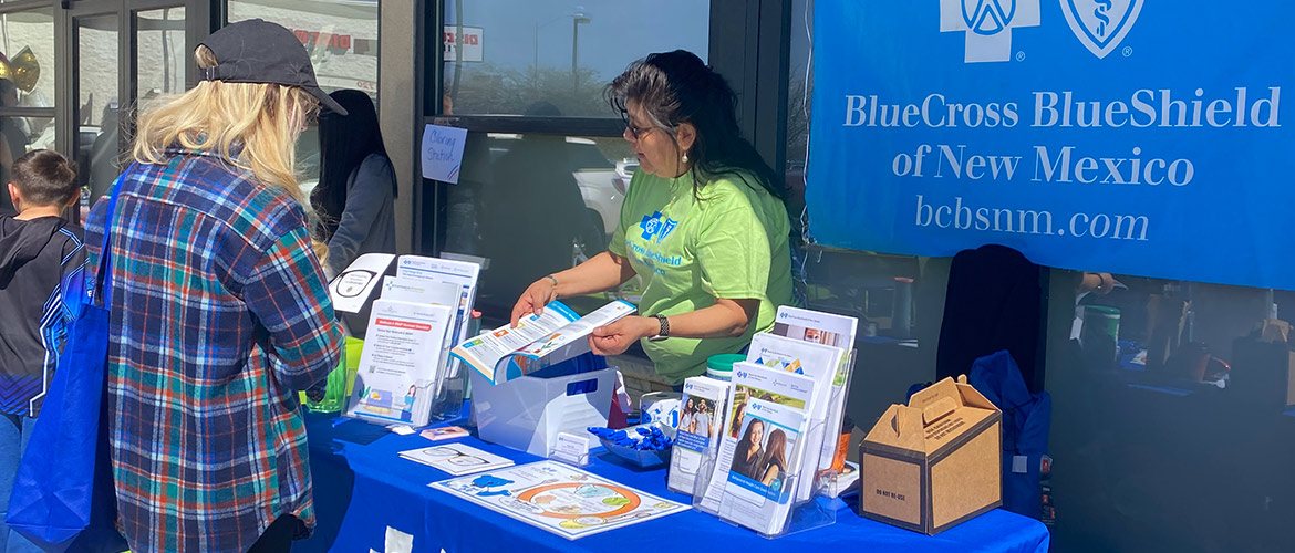 A woman standing behind a Blue Cross and Blue Shield of New Mexico table offers information to someone standing in front of her at an outdoor event